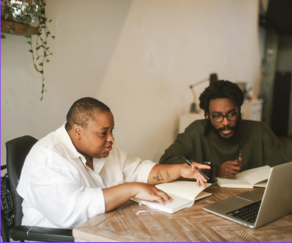 Two people, one in a wheelchair, sitting at a table with a laptop